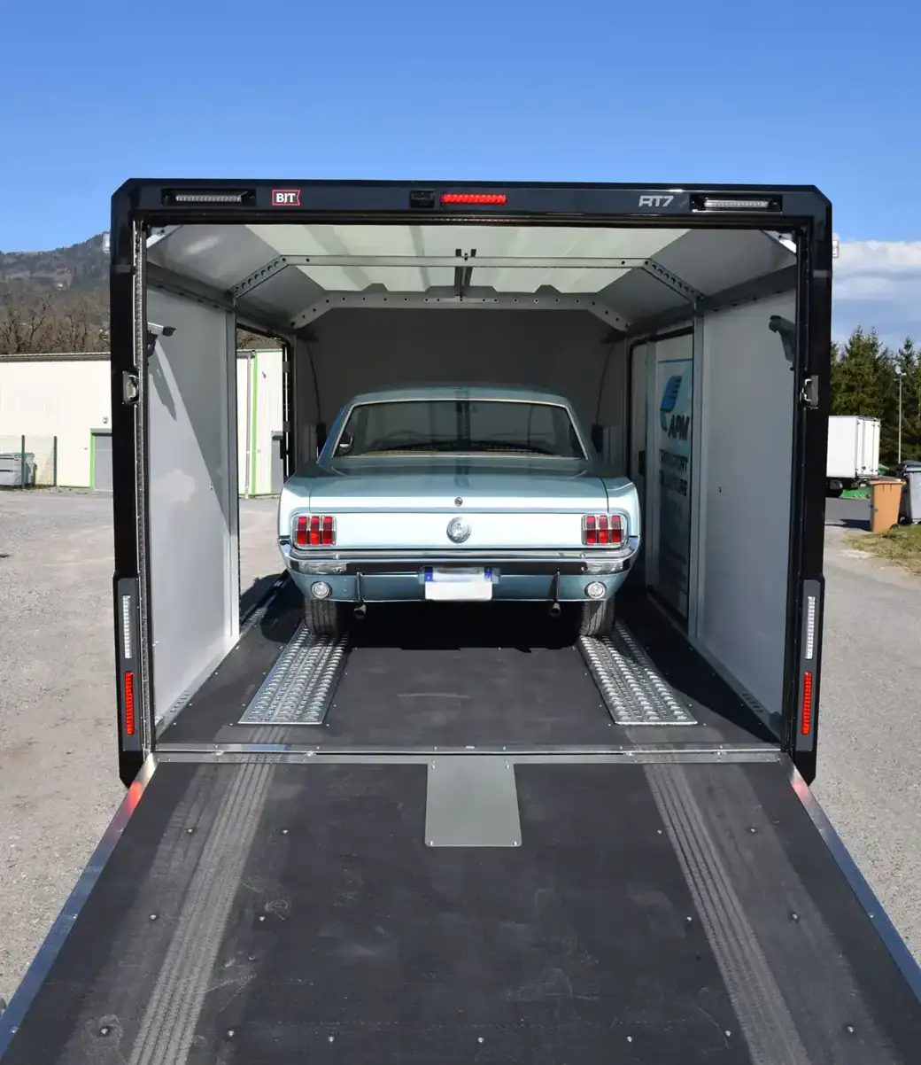 Une voiture ancienne bleu clair garée à l’intérieur d’une remorque noire fermée, la porte arrière abaissée.