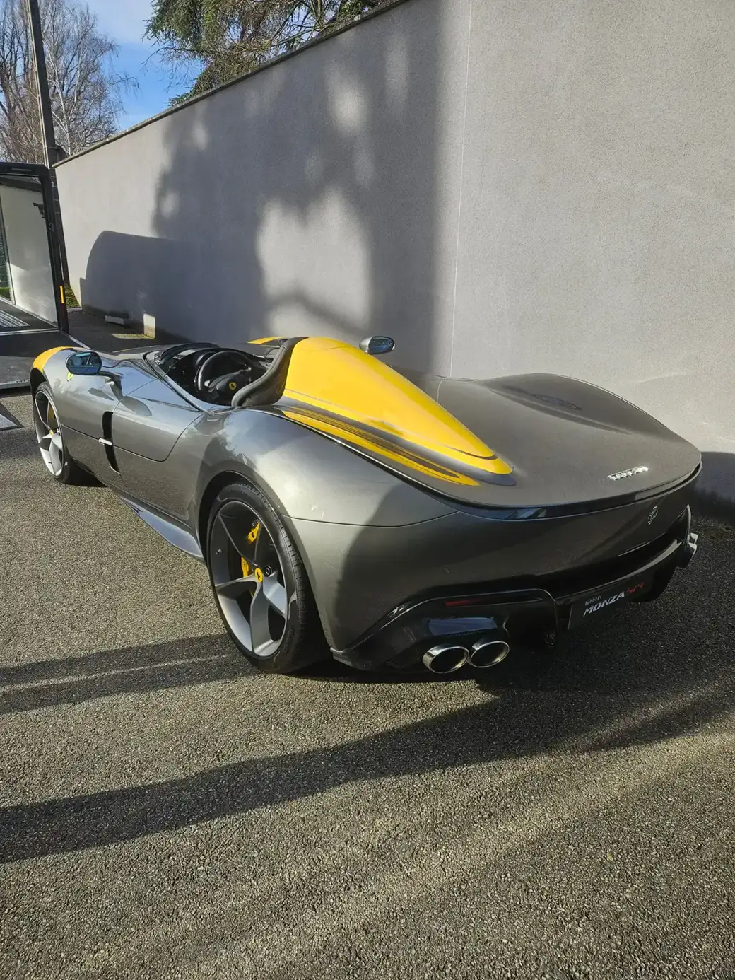 Une supercar Ferrari Monza SP1 grise avec un carénage aérodynamique jaune, garée à l’extérieur sur une surface gravillonnée.