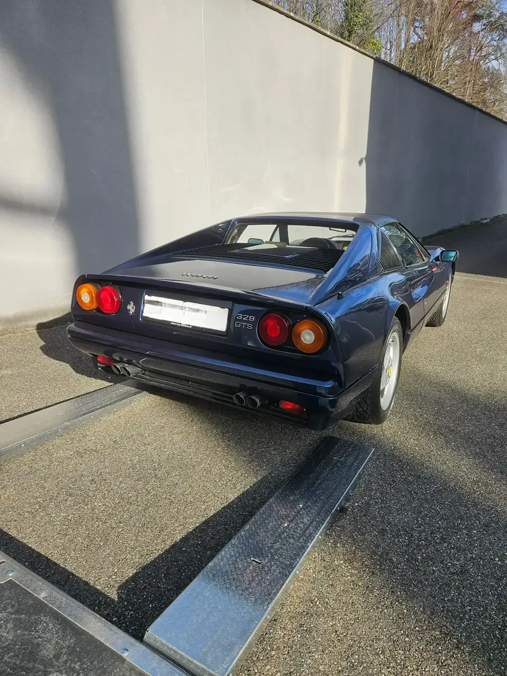 Une Ferrari vintage bleu foncé, garée sur une allée de gravier à côté d’un mur en béton.