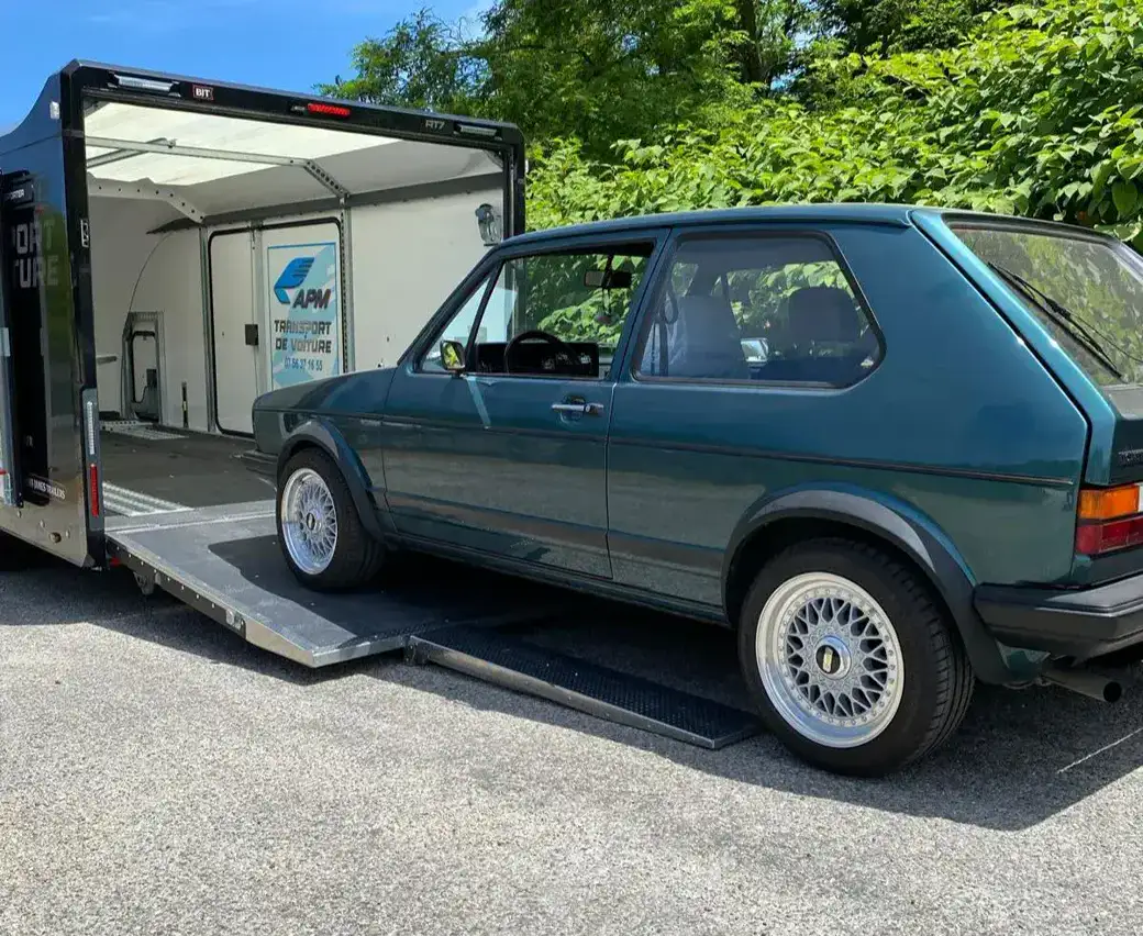 Une Volkswagen Golf vintage turquoise garée sur la rampe métallique d’une remorque porte-voitures noire ouverte.