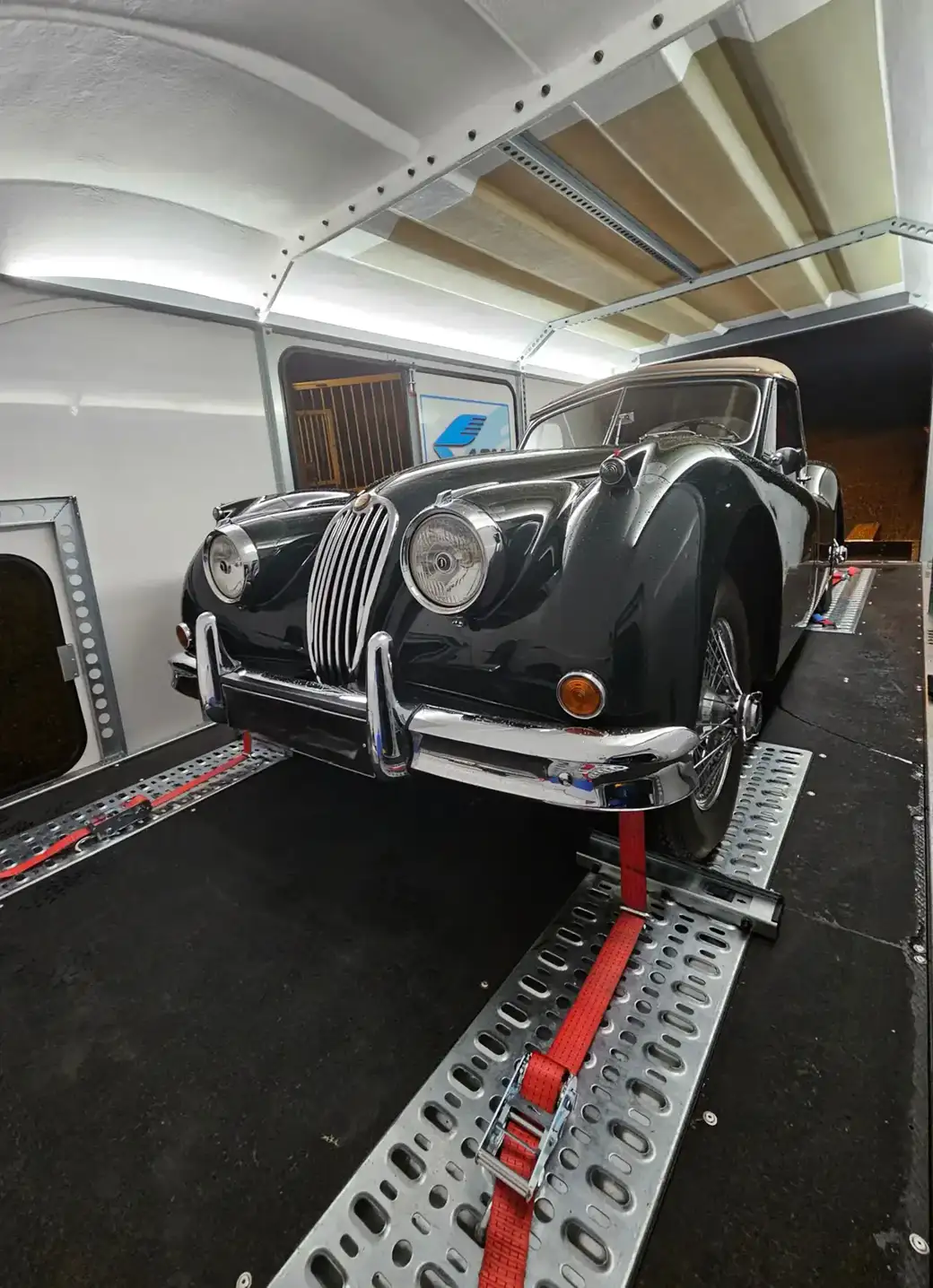 Une Jaguar vintage noire, arrimée par des sangles rouges à l’intérieur d’une remorque blanche.