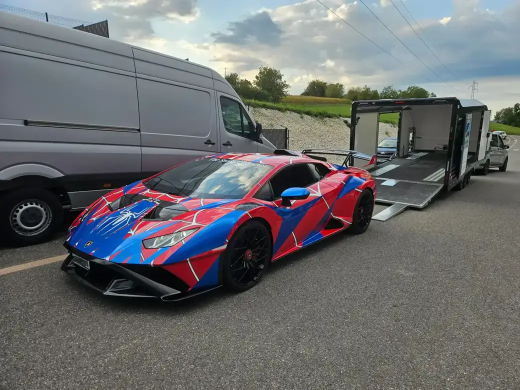 Une Lamborghini rouge et bleue aux couleurs de Spider-Man est garée à côté d’une grande remorque et d’une camionnette.