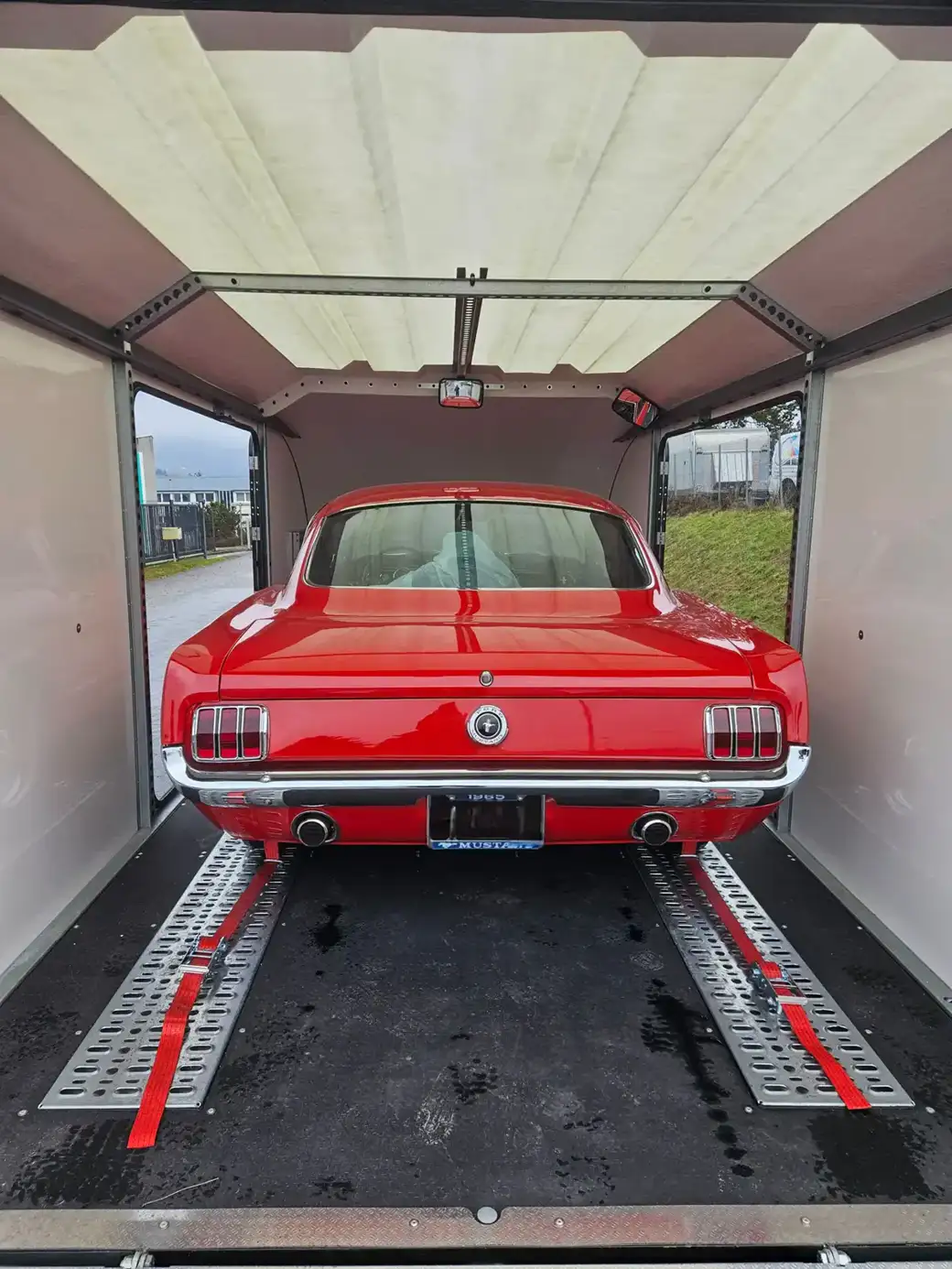 Une Ford Mustang classique rouge vif, garée à l’intérieur d’une remorque fermée et arrimée au sol par des sangles rouges.