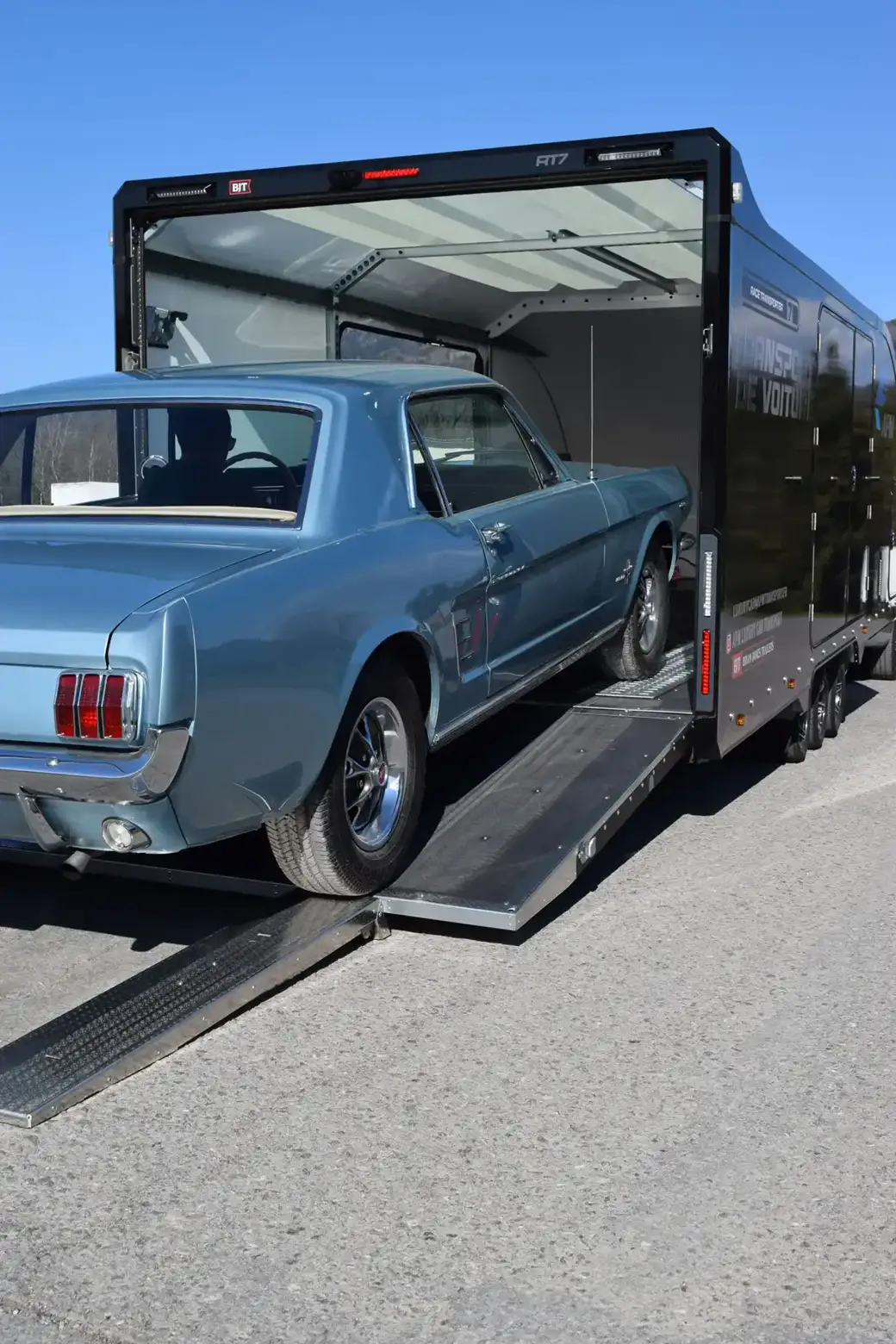 Une Ford Mustang vintage bleu clair est chargée sur une remorque fermée noire à l’aide d’une rampe.