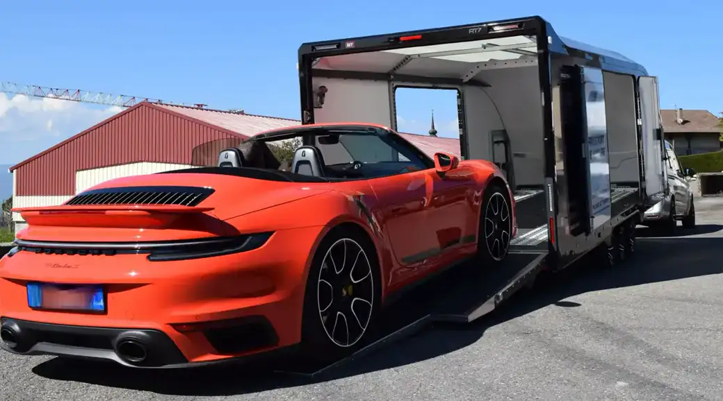 Une Porsche 911 cabriolet orange descend la rampe d’une remorque porte-voitures fermée.