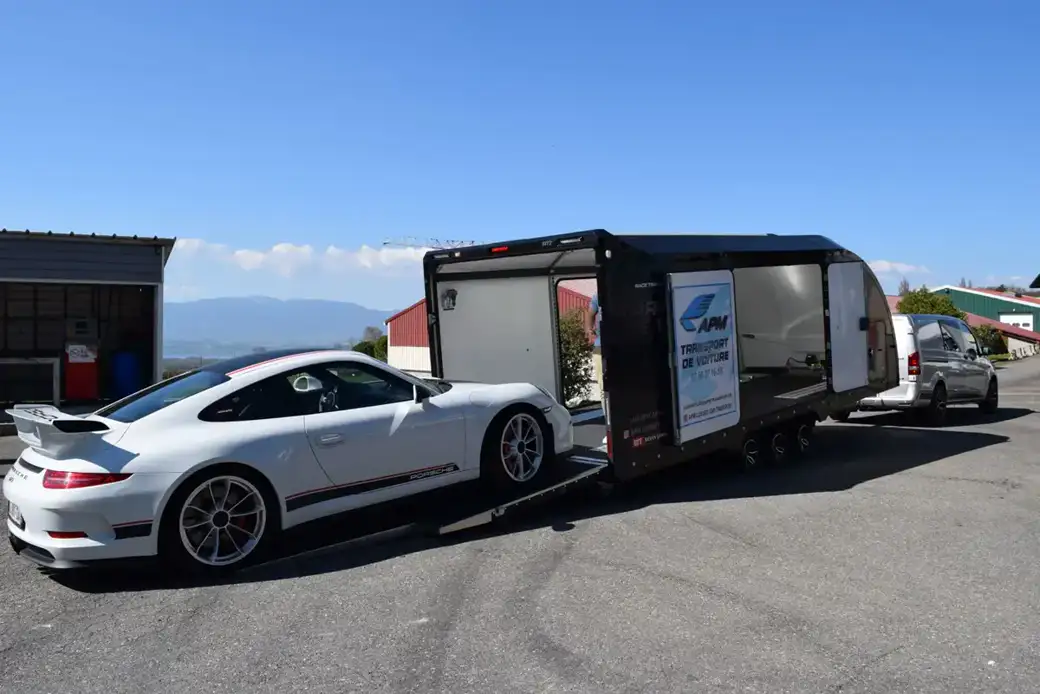 Une Porsche blanche est chargée sur une remorque porte-voitures ouverte, sur un parking extérieur goudronné.