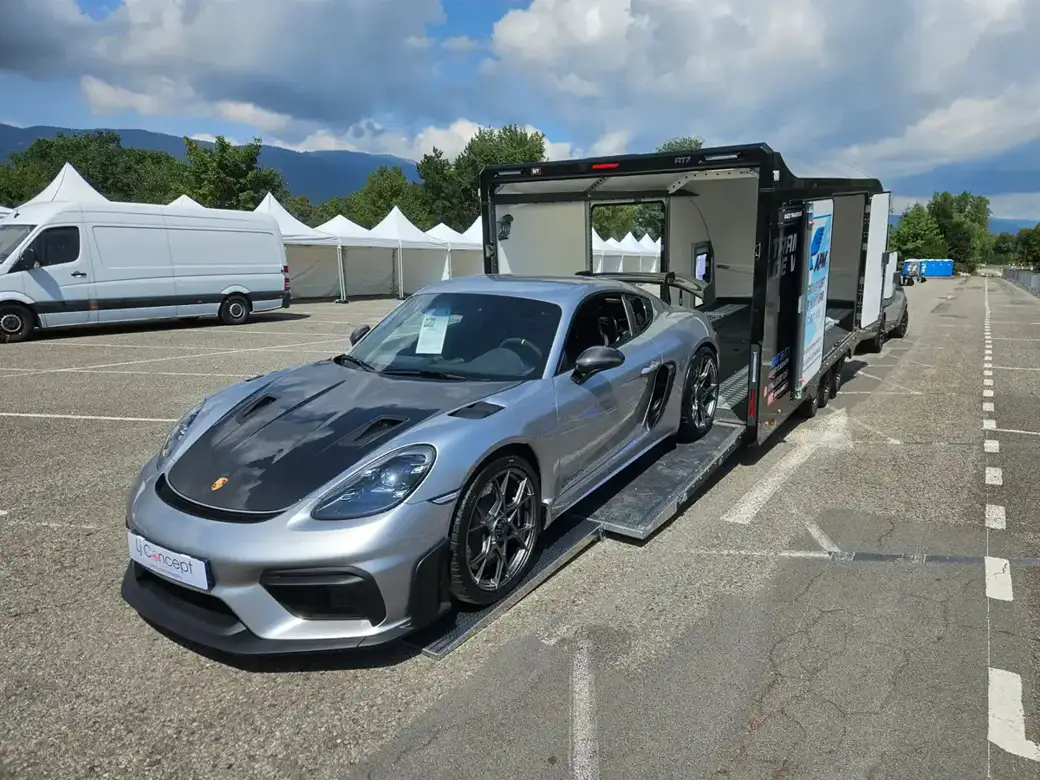 Une Porsche 718 Cayman GT4 RS argentée, chargée sur la rampe d’une remorque porte-voitures fermée.