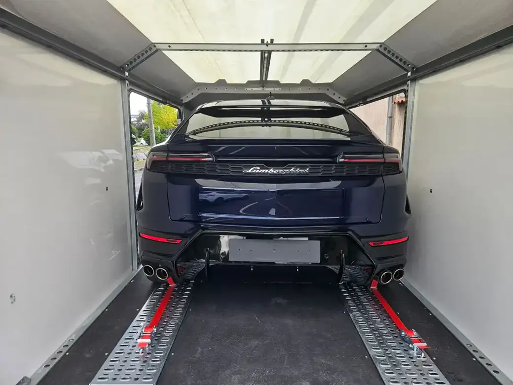 Un SUV Lamborghini Urus bleu foncé, arrimé par des sangles rouges à l’intérieur d’une remorque porte-voitures fermée.